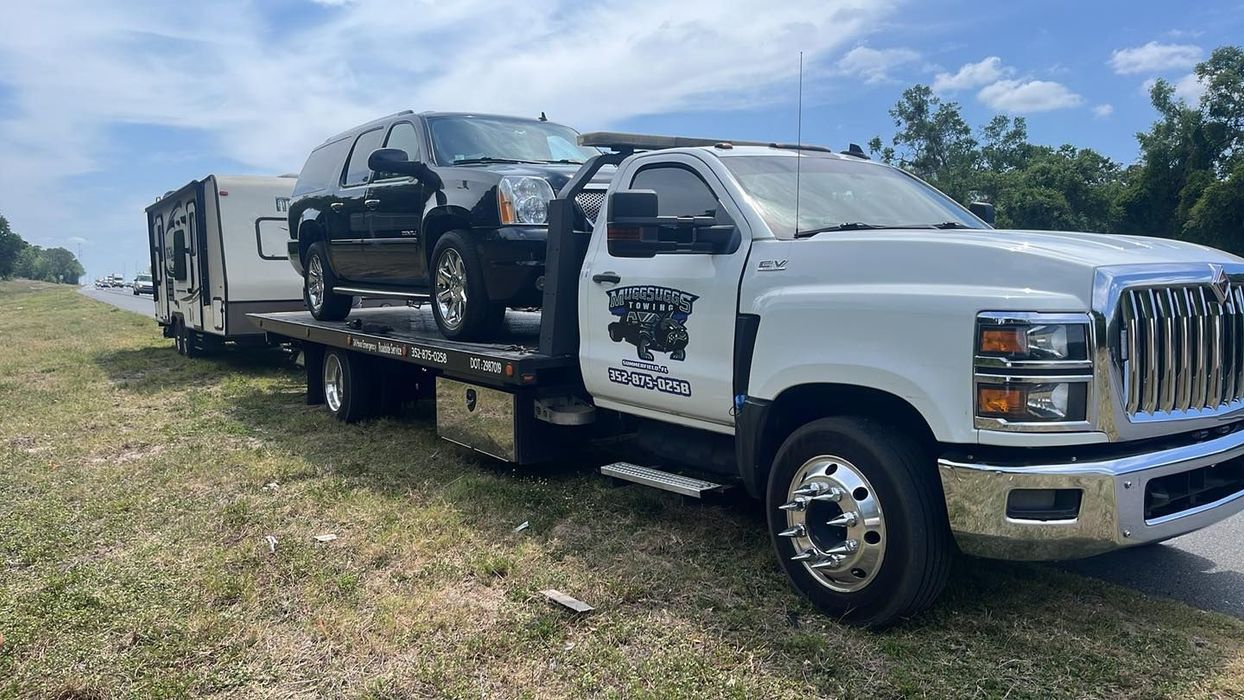 A white tow truck carrying a black truck and camper trailer on a highway, sunny day.