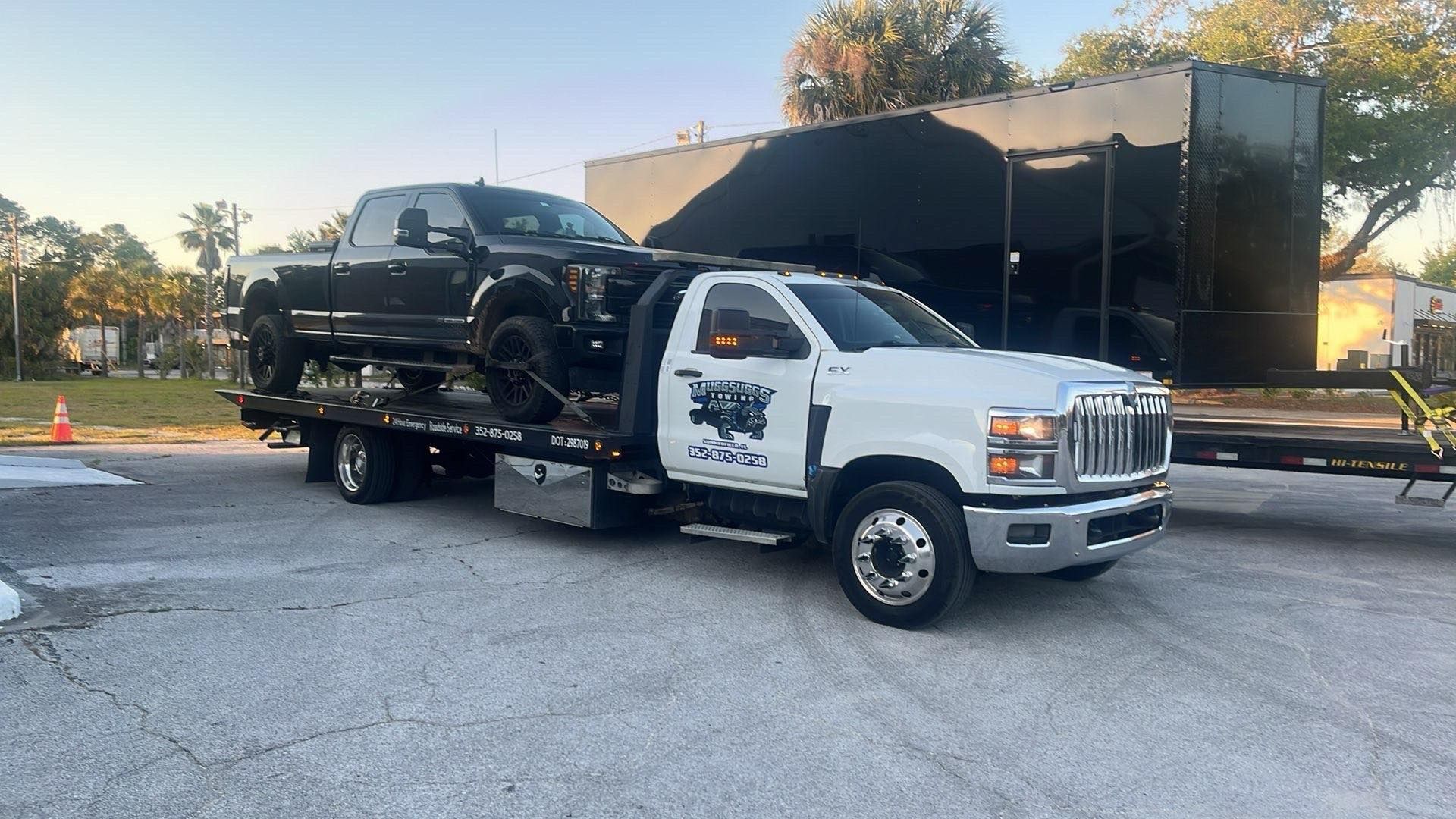 White tow truck with a dark pickup truck loaded on the flatbed, parked in front of a black trailer.