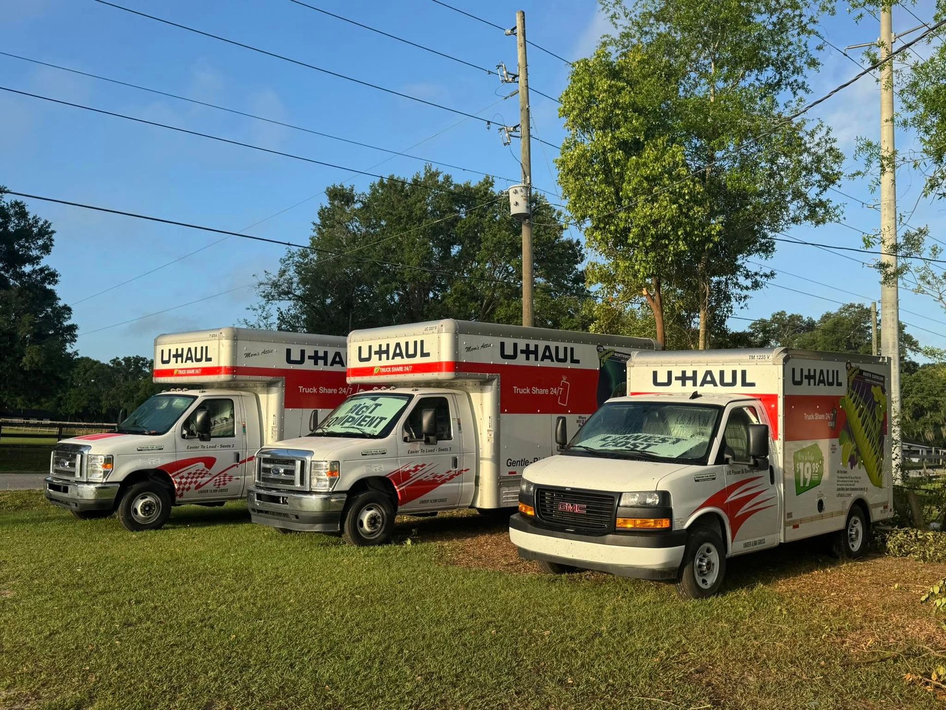 Three U-Haul trucks parked on a grassy area with trees and power lines in the background.