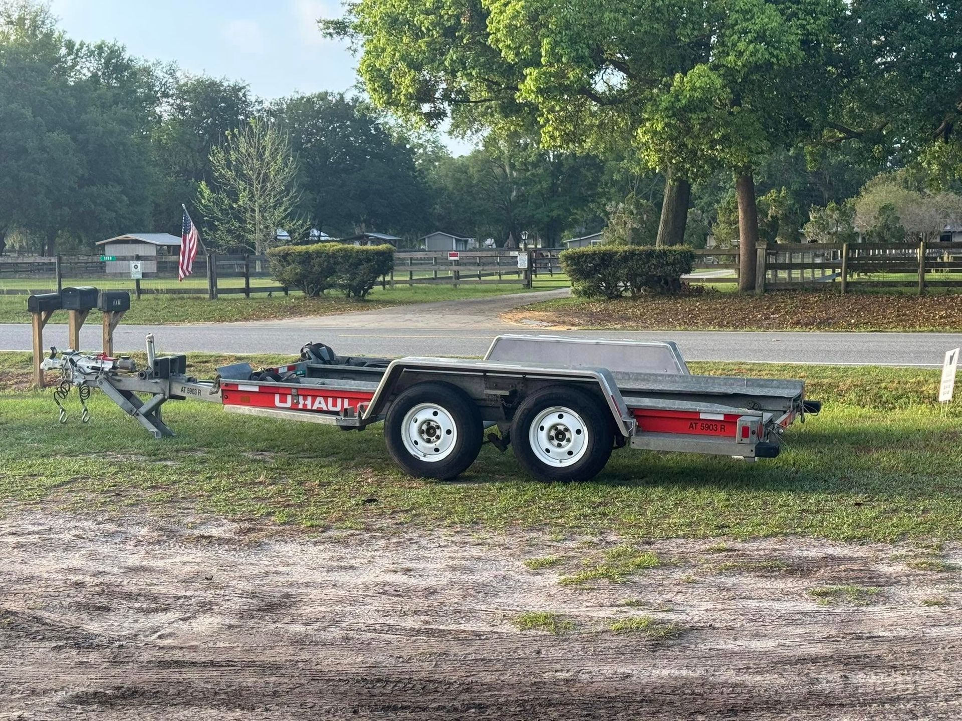 U-Haul car trailer parked on grass, in front of a fence and trees.