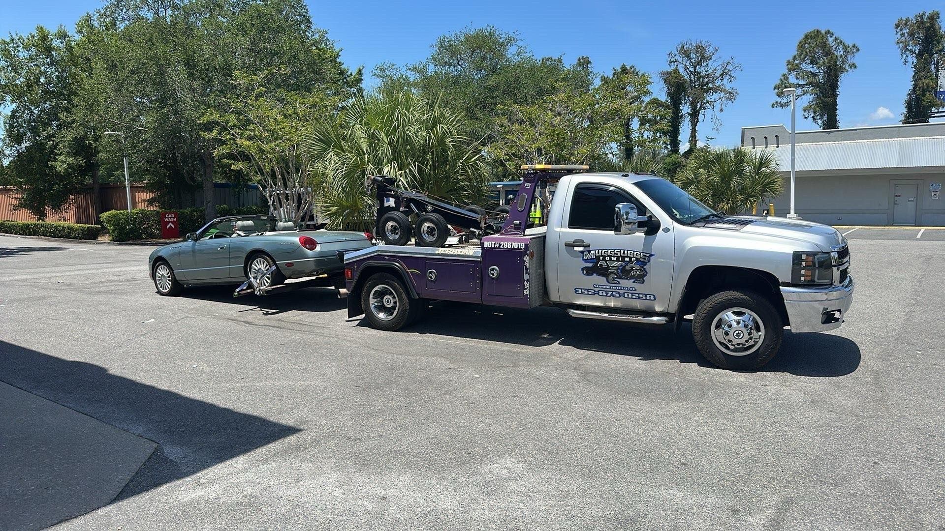 Silver tow truck towing a silver convertible car in a parking lot on a sunny day.