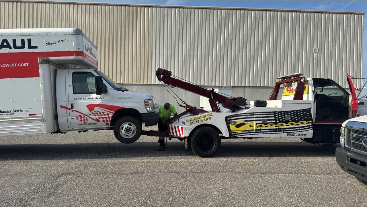 A U-Haul truck being towed by a tow truck, likely due to a breakdown, outdoors, bright sunny day.