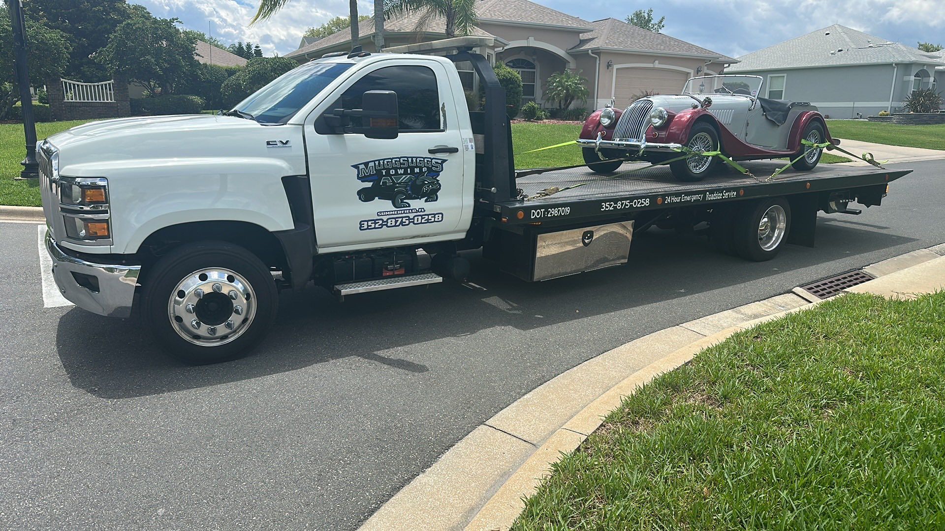 White tow truck hauling a classic red convertible on a sunny residential street.
