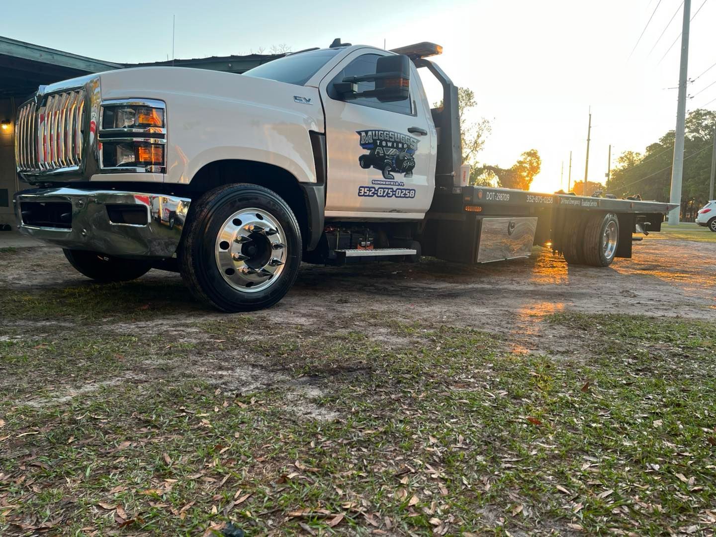 White tow truck parked on grass, with sun setting in the background.
