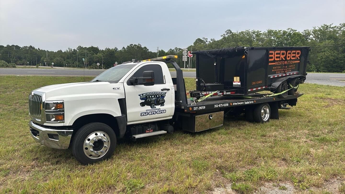 White truck with a black dumpster on a flatbed trailer, parked on a grassy area near a road.