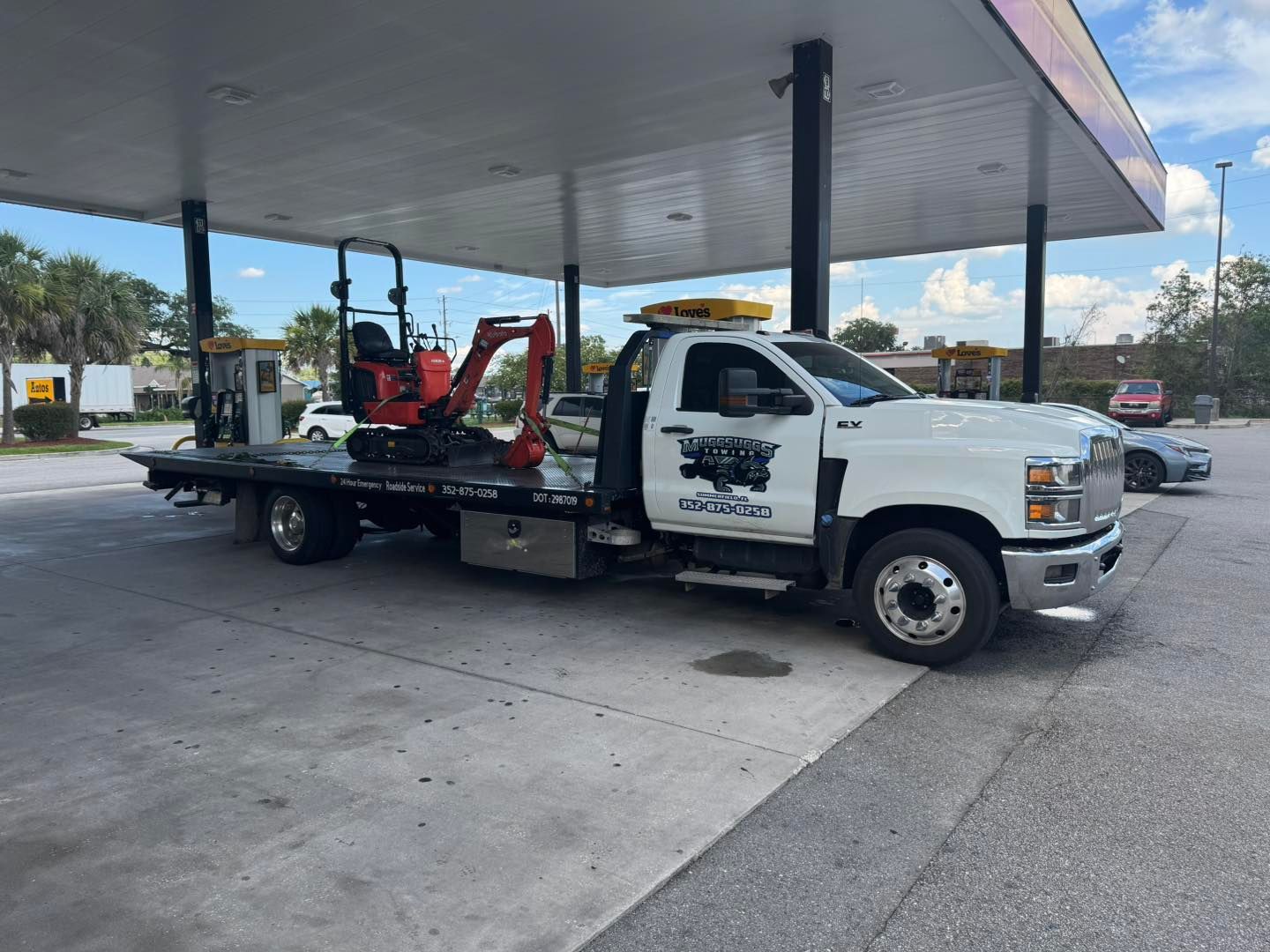 White tow truck at a gas station, carrying a small orange excavator.