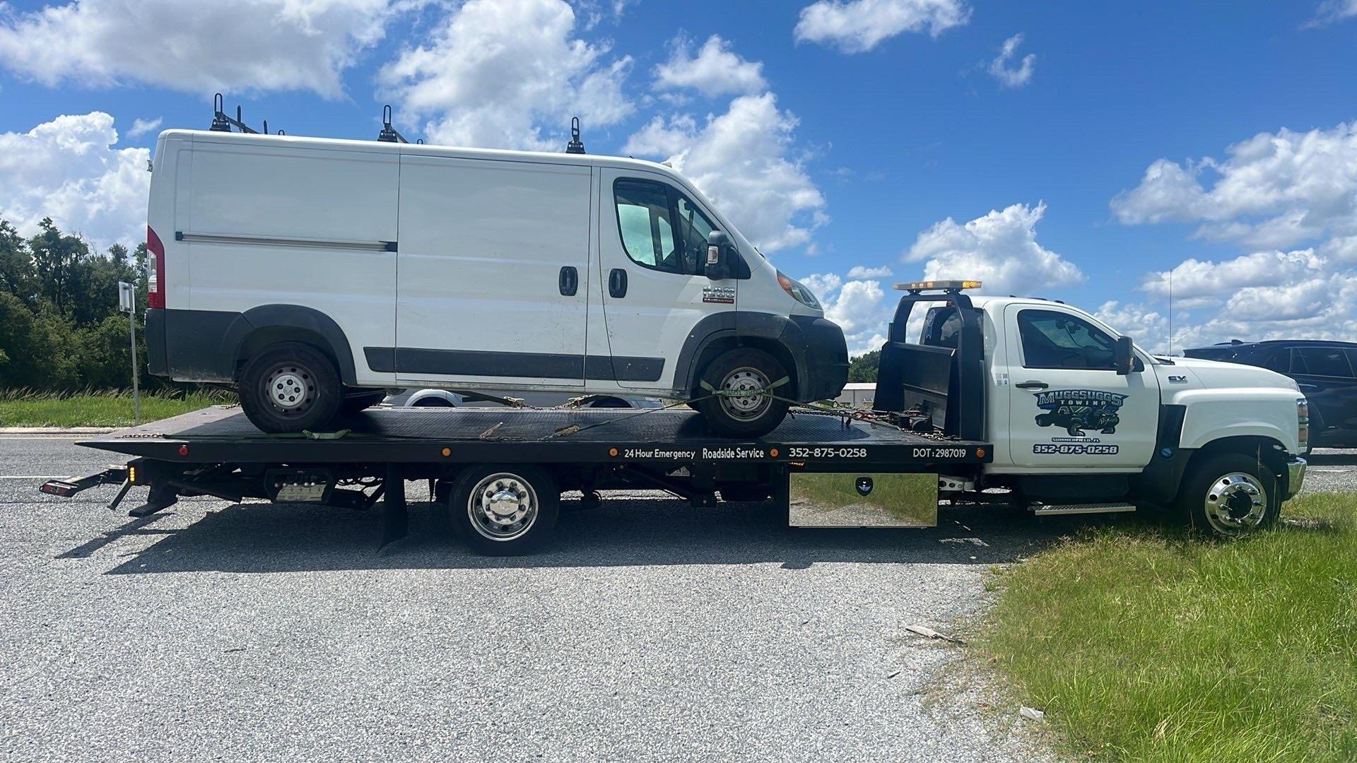 White van on a flatbed tow truck on a gravel road, under a blue sky with clouds.