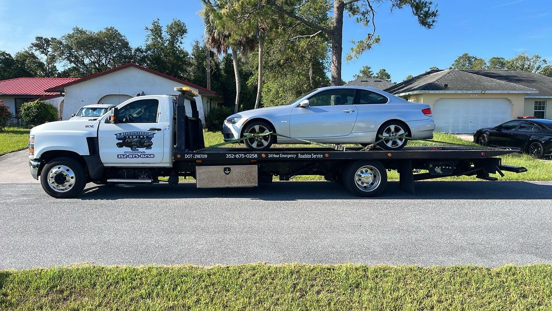 Tow truck carrying a silver sedan on a residential street under a blue sky.
