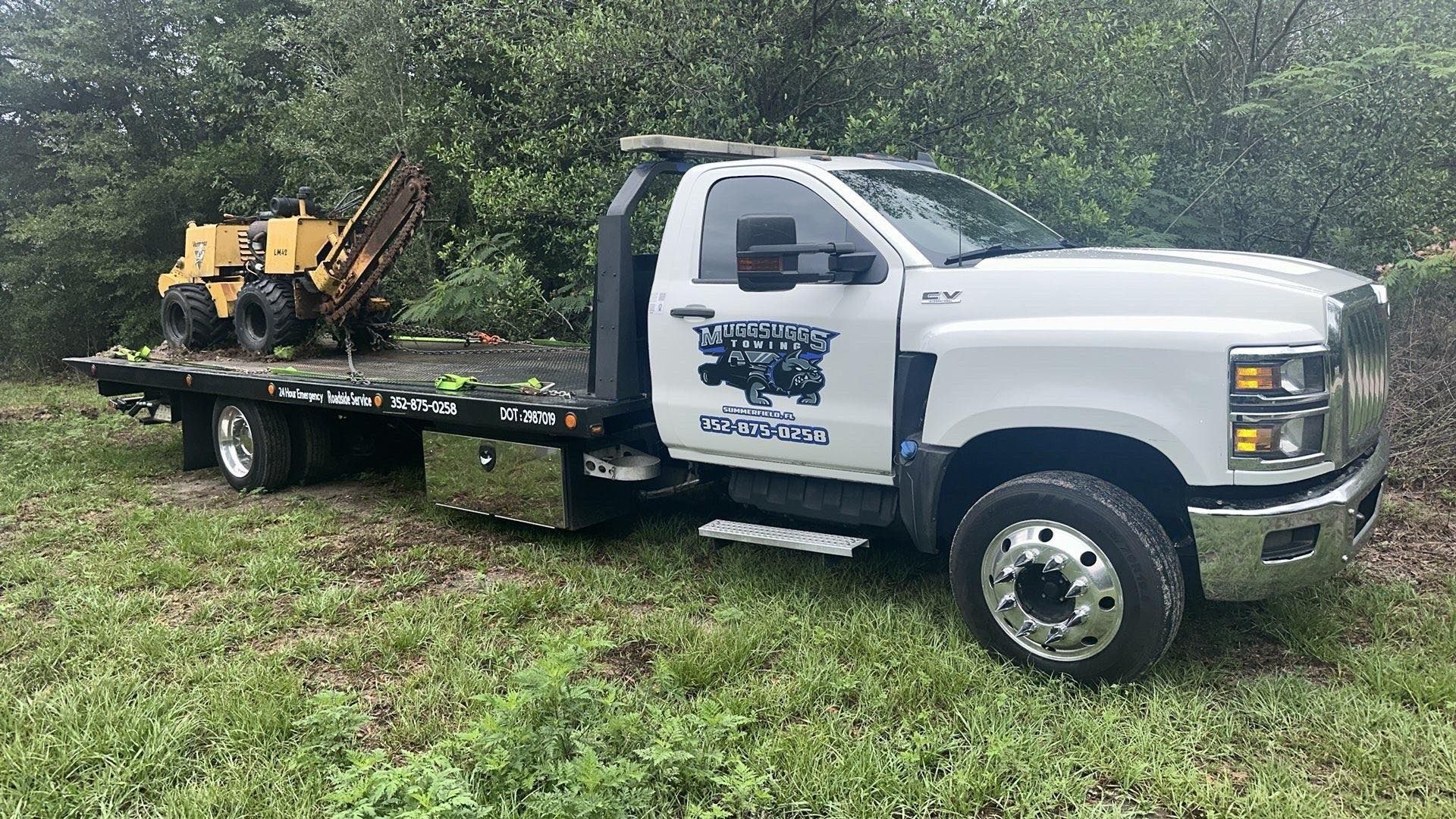 White tow truck with a yellow stump grinder on a grassy area near trees.