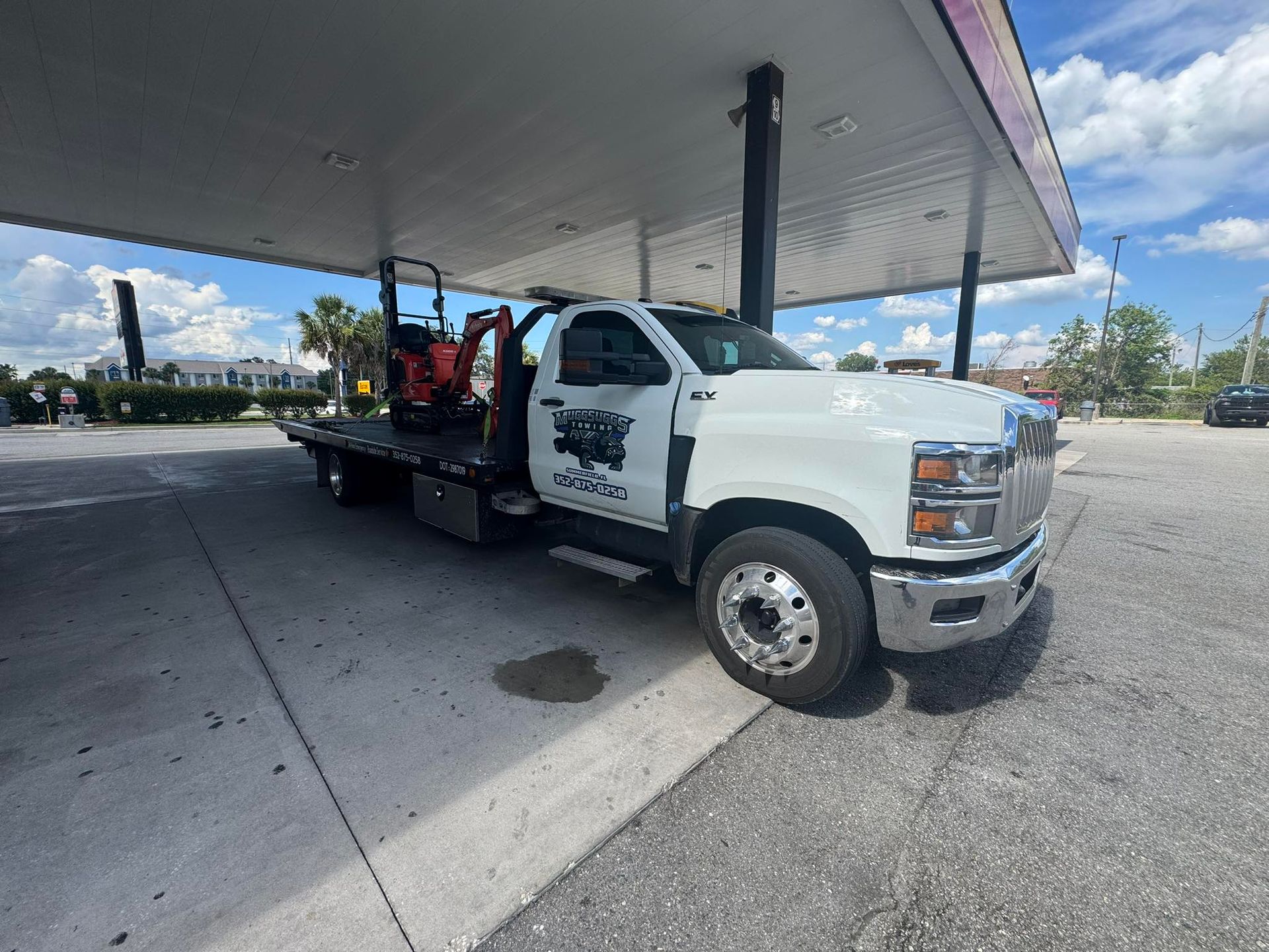 White flatbed truck with an excavator at a gas station on a sunny day.