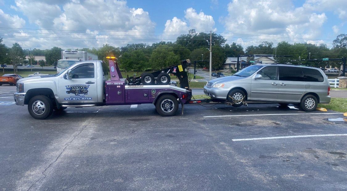 A tow truck hauling a silver minivan in a parking lot on a cloudy day.