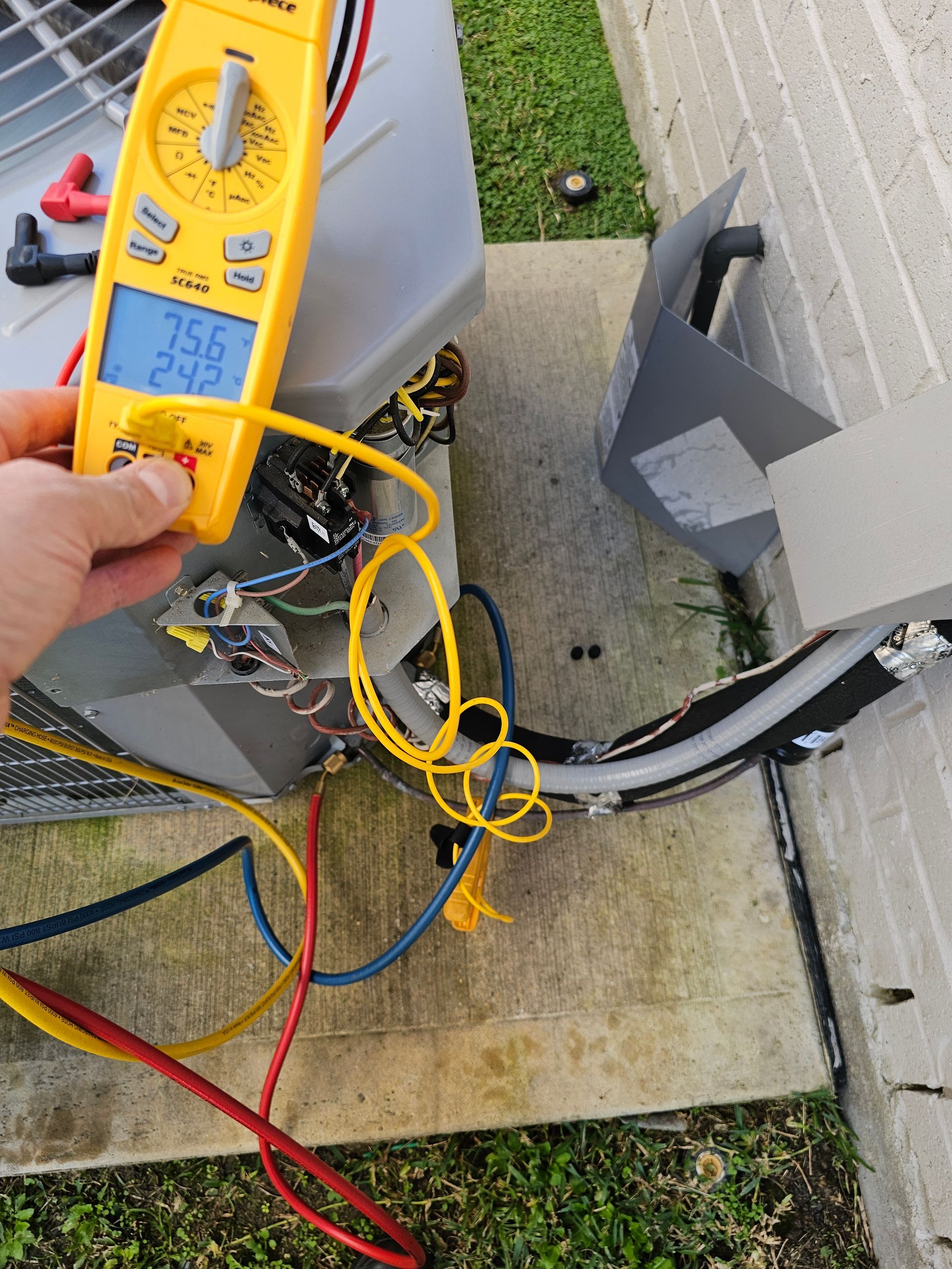 A person is holding a digital multimeter next to an air conditioner.