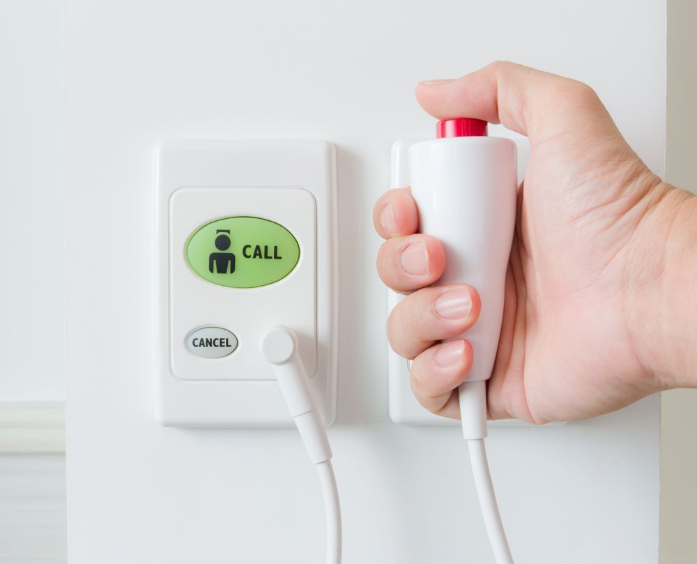Hand pressing a red emergency button connected to a white wall-mounted call system.