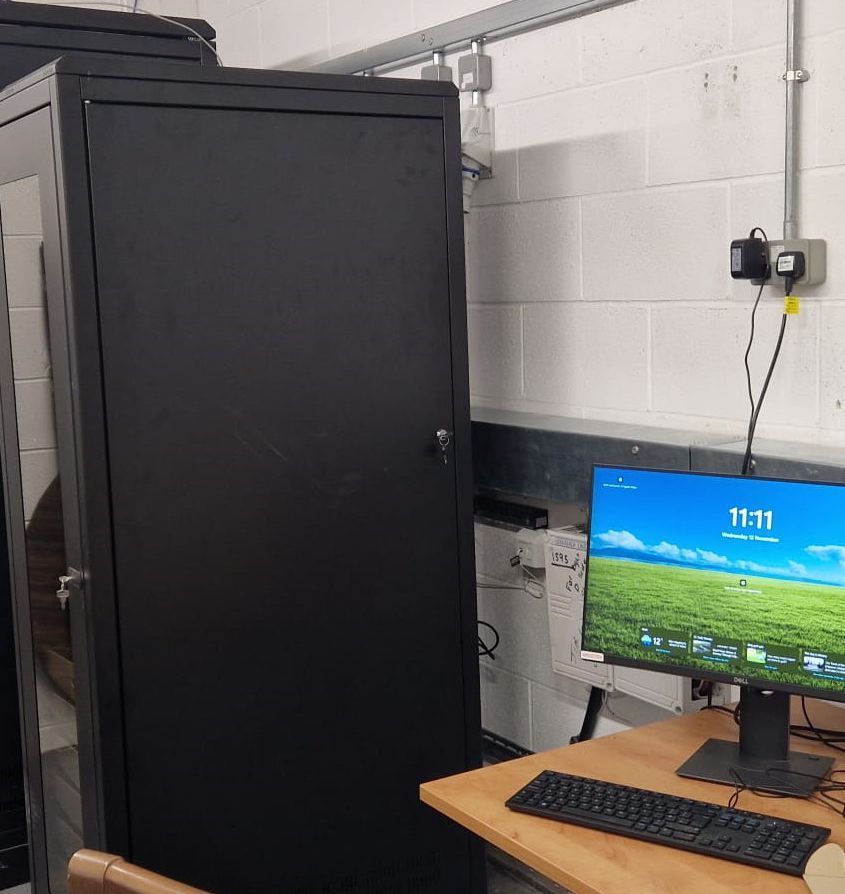 Black server rack next to a desk with a computer and keyboard in a room with white walls.