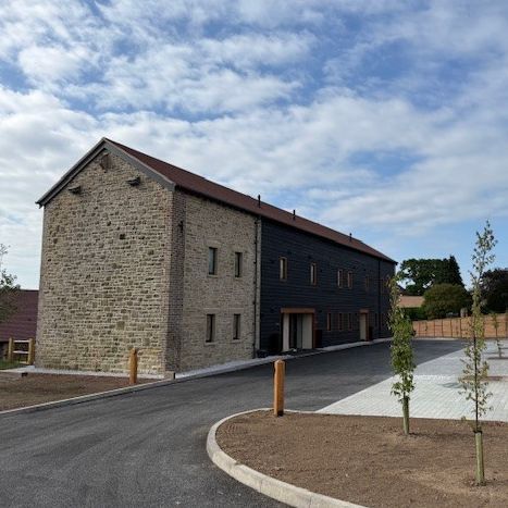 Stone and black-sided building with dark roof, driveway, and small trees on a cloudy day.