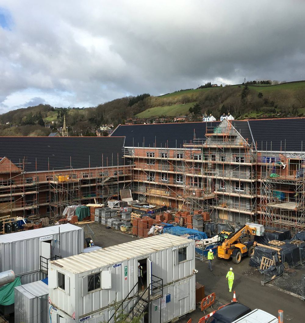 Construction site with partially built brick buildings, scaffolding, and storage containers under a cloudy sky.