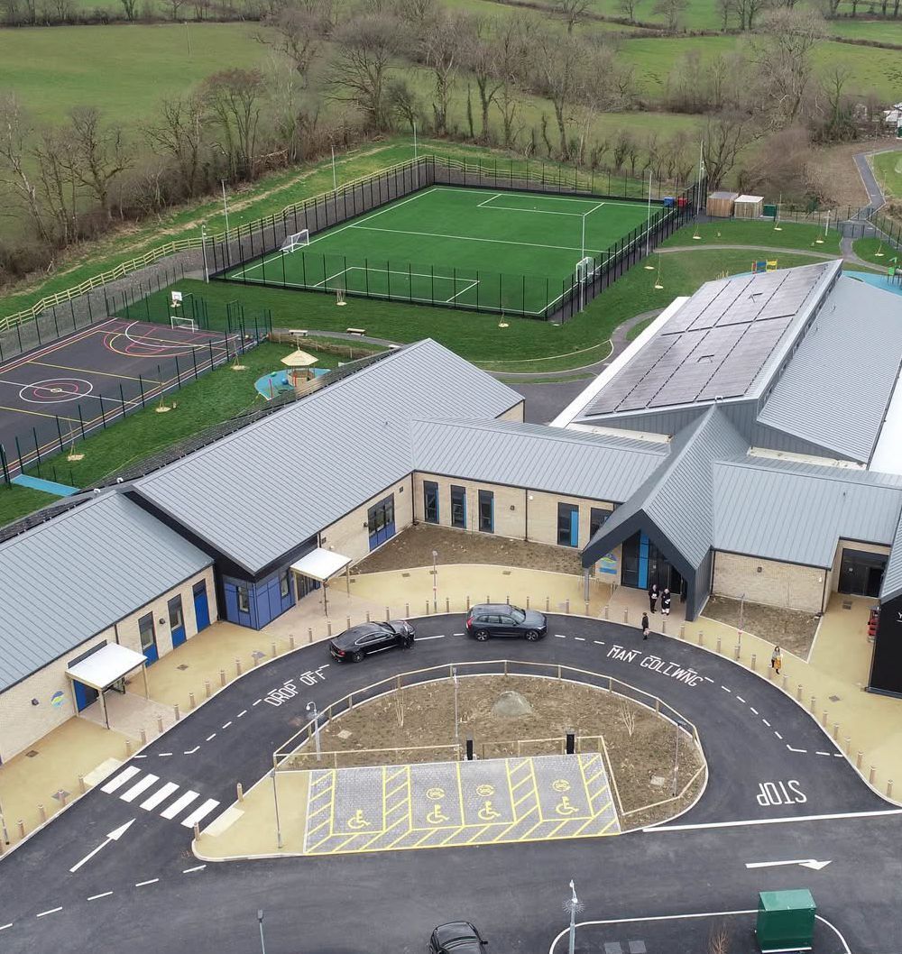 Aerial view of a school building with a sports field, playground, and circular driveway with two cars parked.