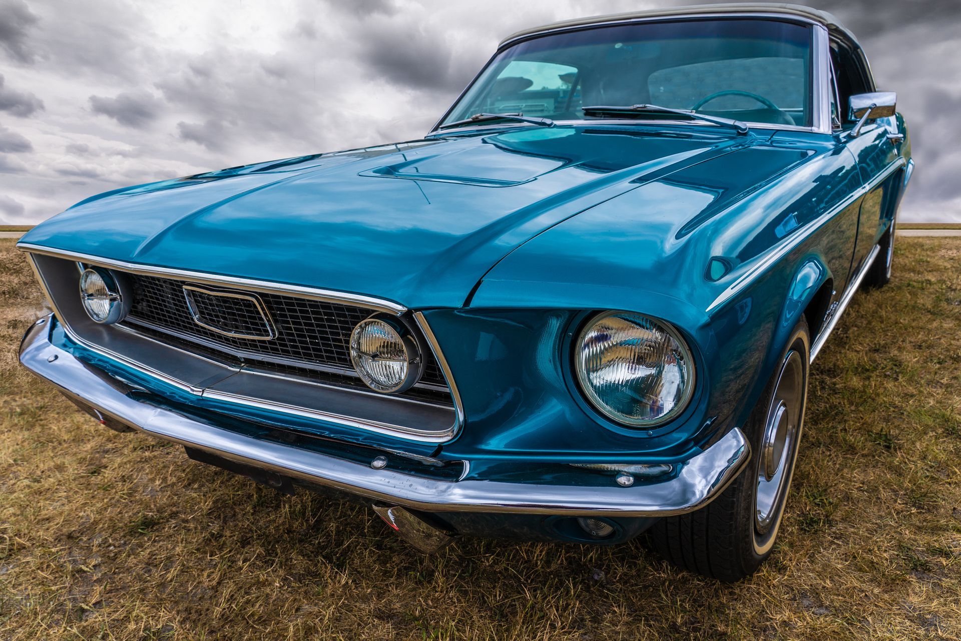 A blue mustang is parked in a field with a cloudy sky in the background