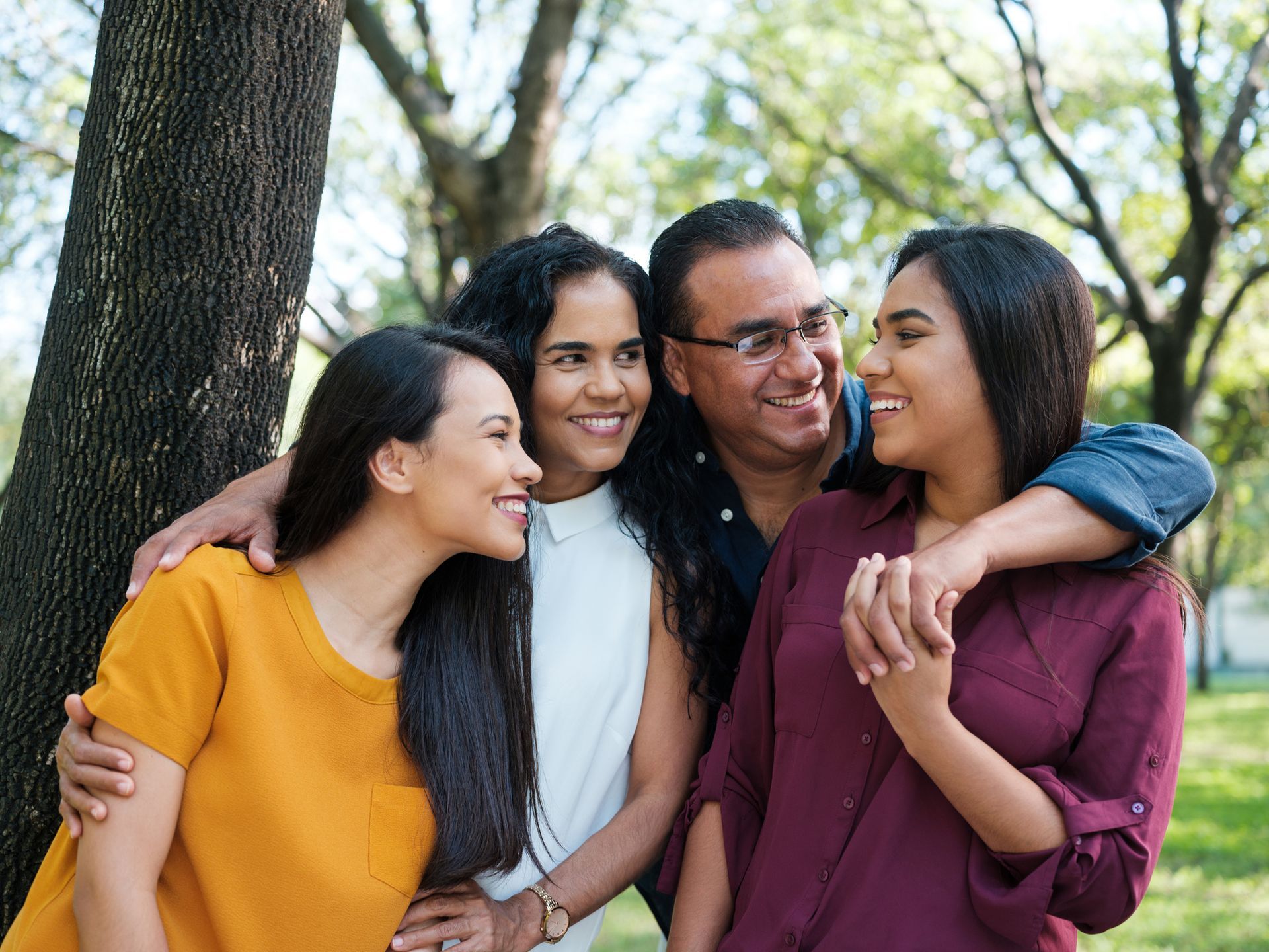 Family of four smiling, hugging in a park next to a tree.