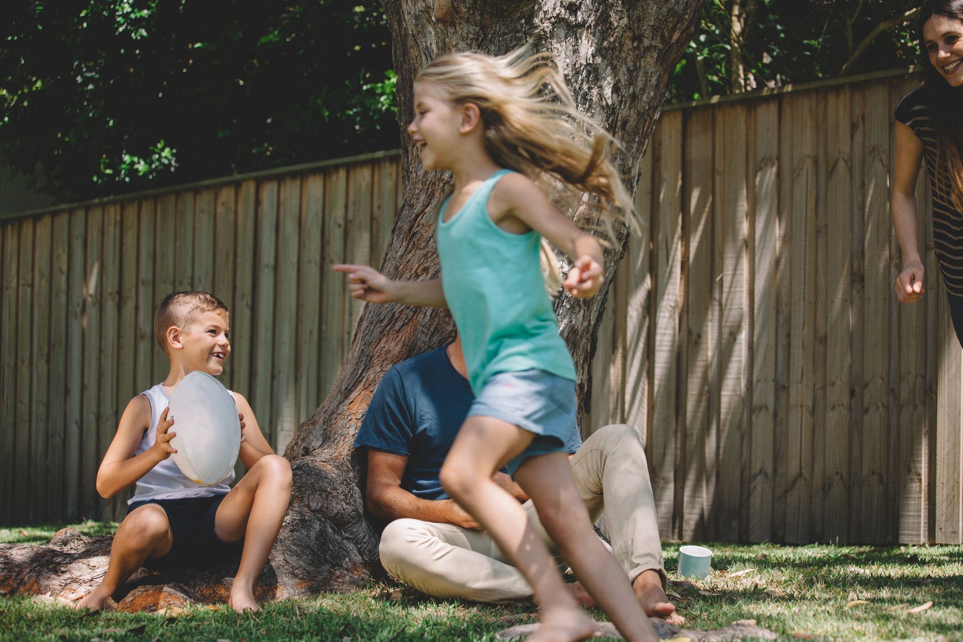 Children playing in a backyard: Girl running, boy holding a frisbee, adult sitting under a tree. Children playing in a backyard: Girl running, boy holding a frisbee, adult sitting under a tree.