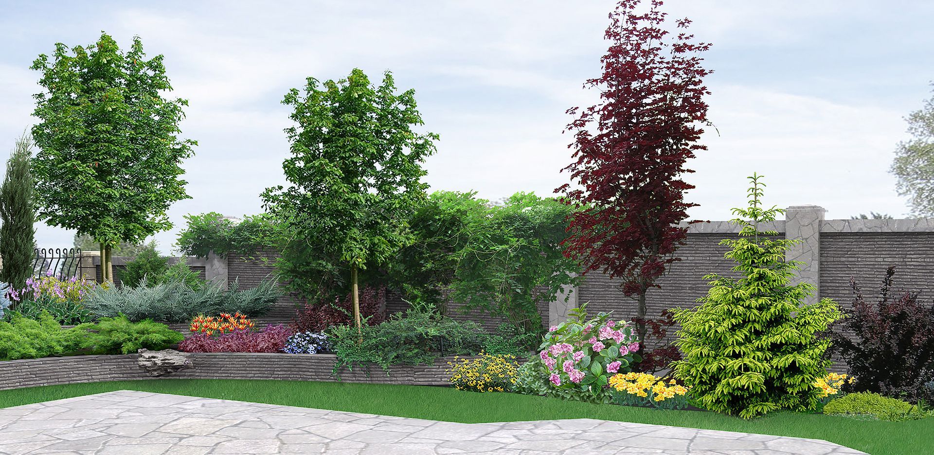 Beautiful landscaped backyard garden with a variety of colorful flowers, shrubs, and trees bordered by a stone fence and paved patio area