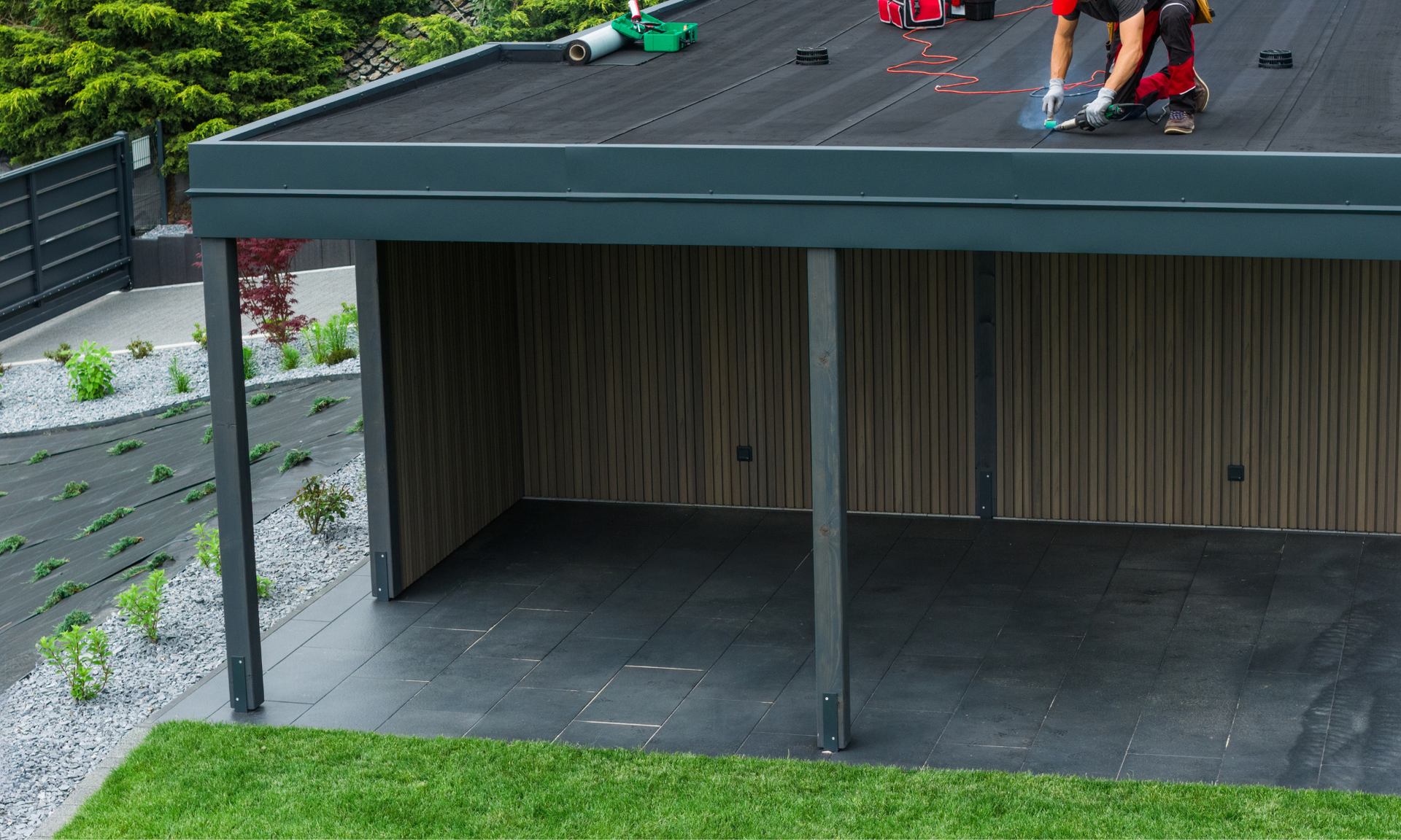 Een persoon werkt op het platte, zwarte dak van een carport. Donkere metalen steunbalken, houten lambrisering, groen gras.