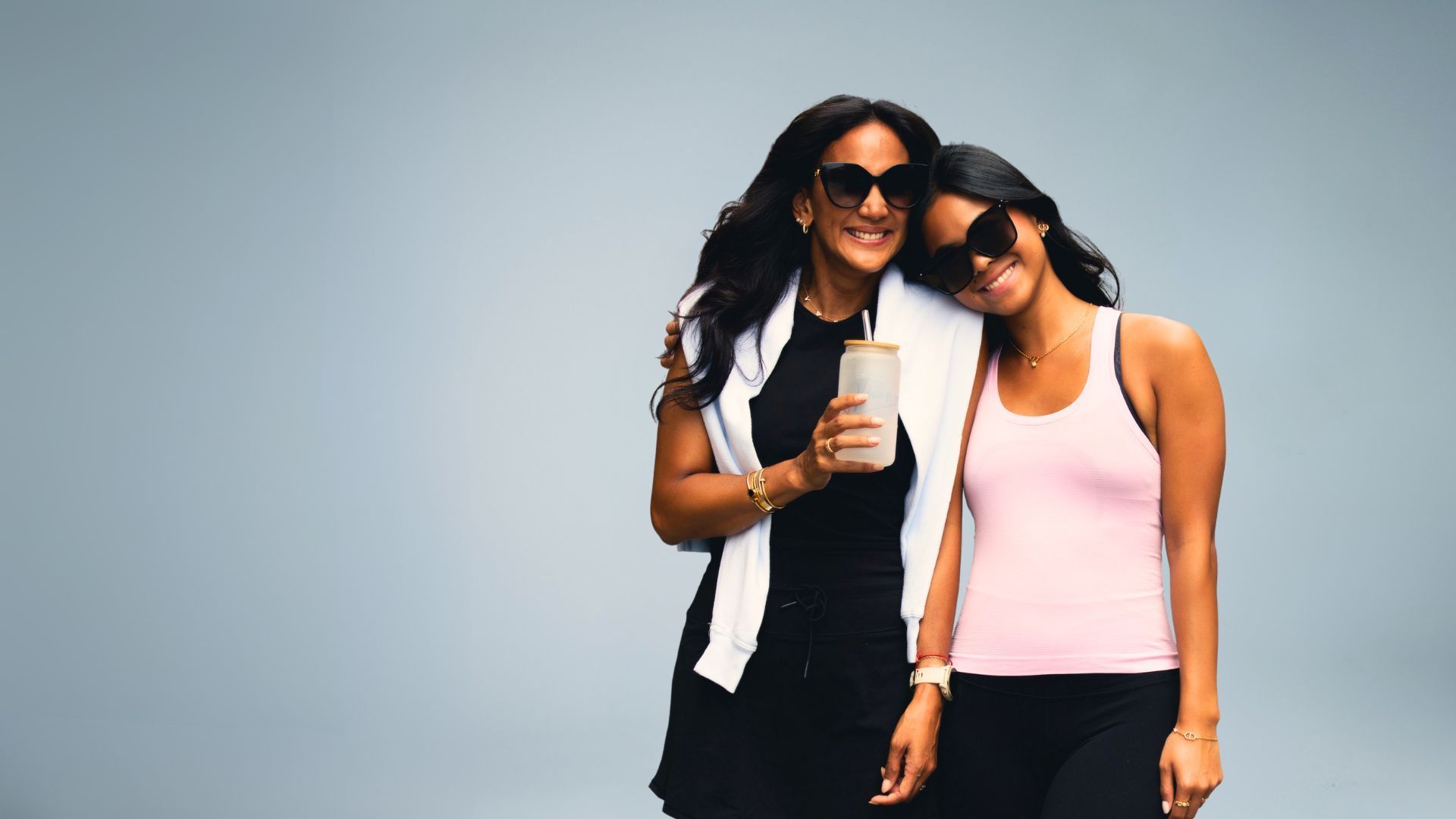 Two women in sunglasses posing together. One holds a drink, the other leans on her shoulder. Against a gray backdrop.