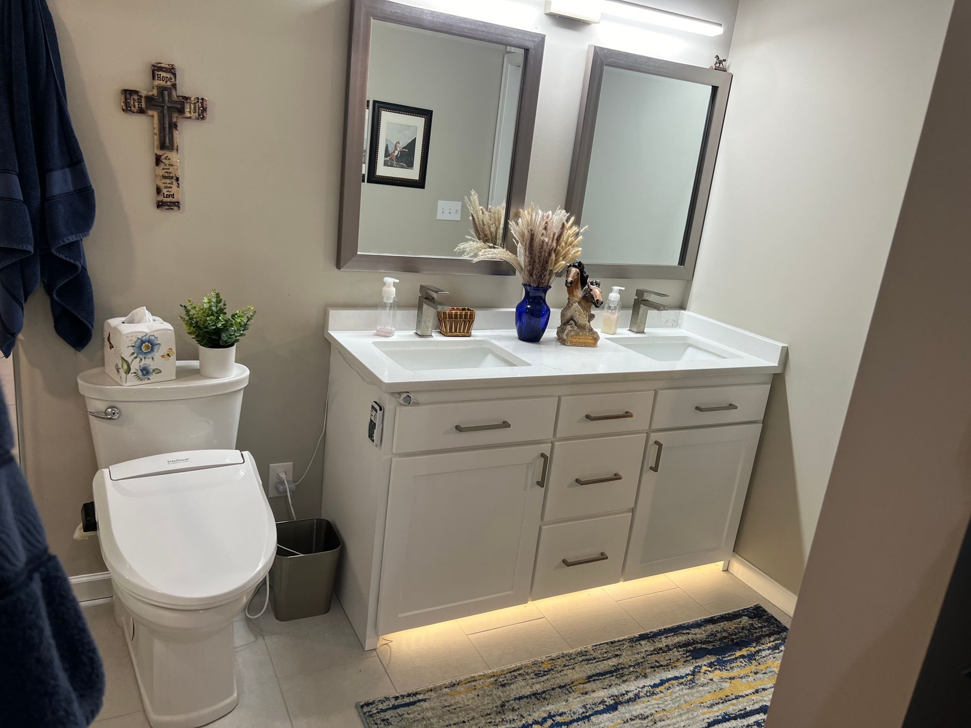 Bathroom with white vanity, mirrors, and toilet. Includes rug, plants, and decorative cross on wall.