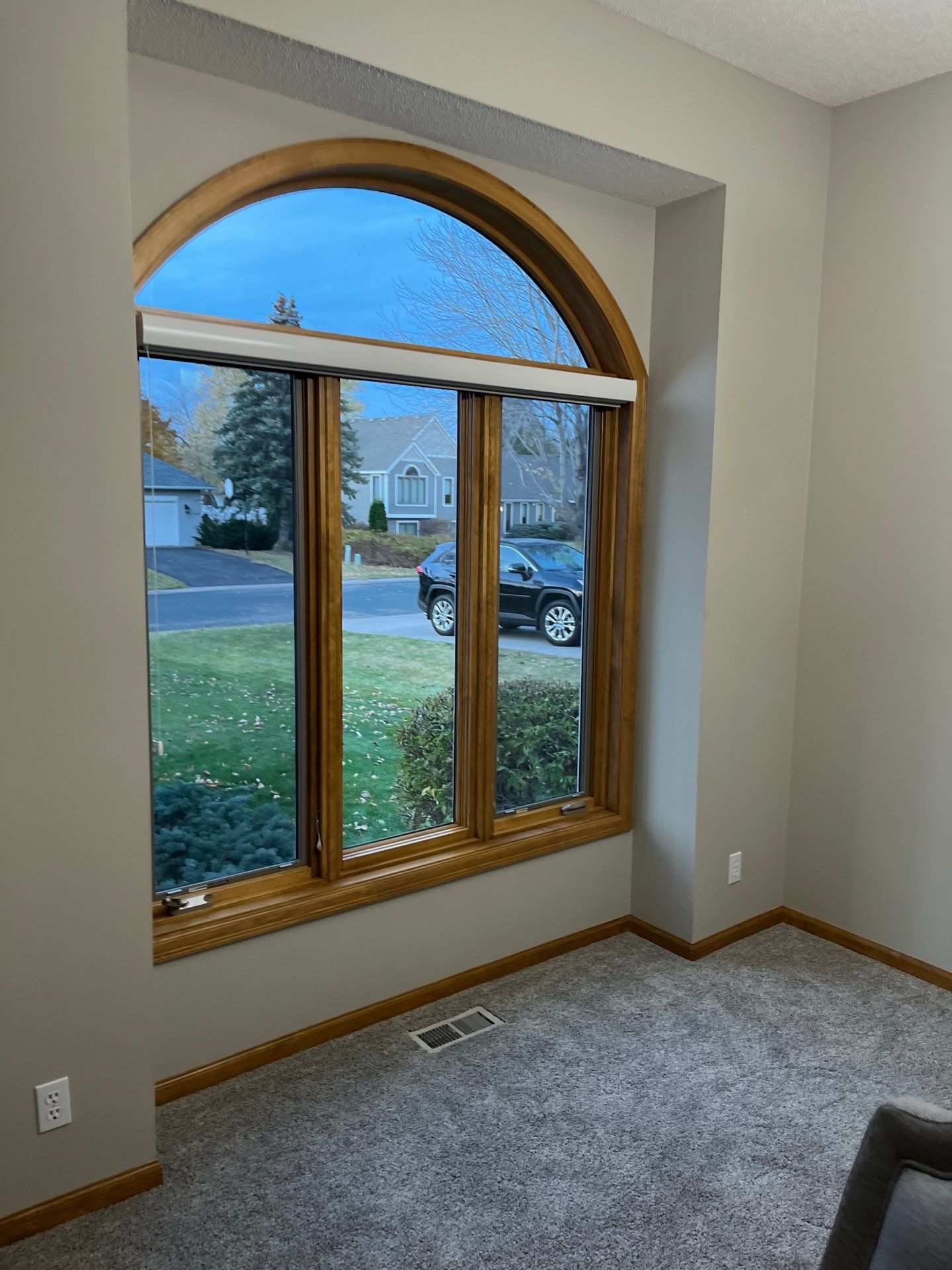 Room with large arched window, brown trim, gray walls, and carpet; view of neighborhood street.