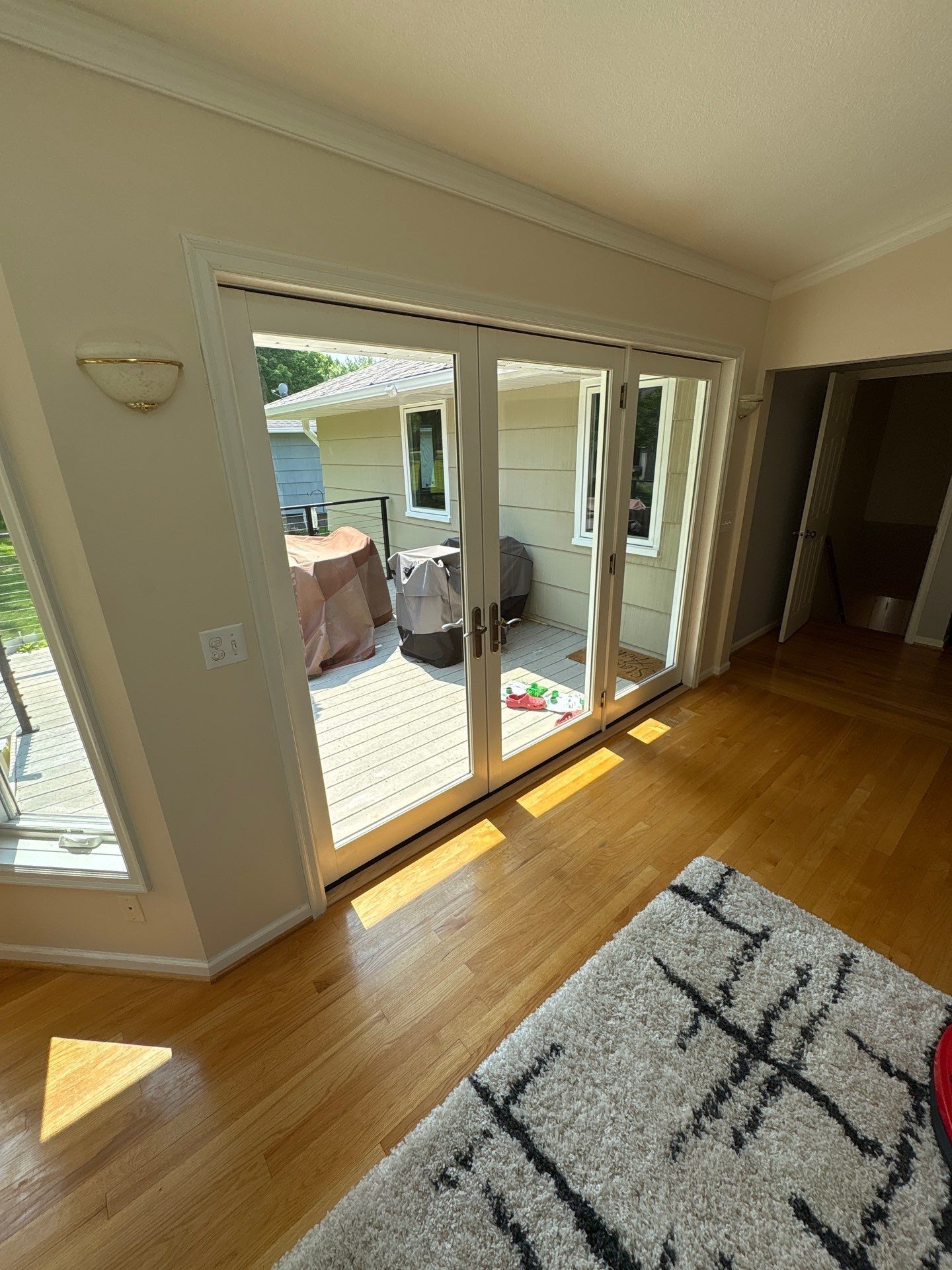 Bright room with patio doors, wooden floors, beige walls, and a gray rug. View of outdoor deck with covered grill.
