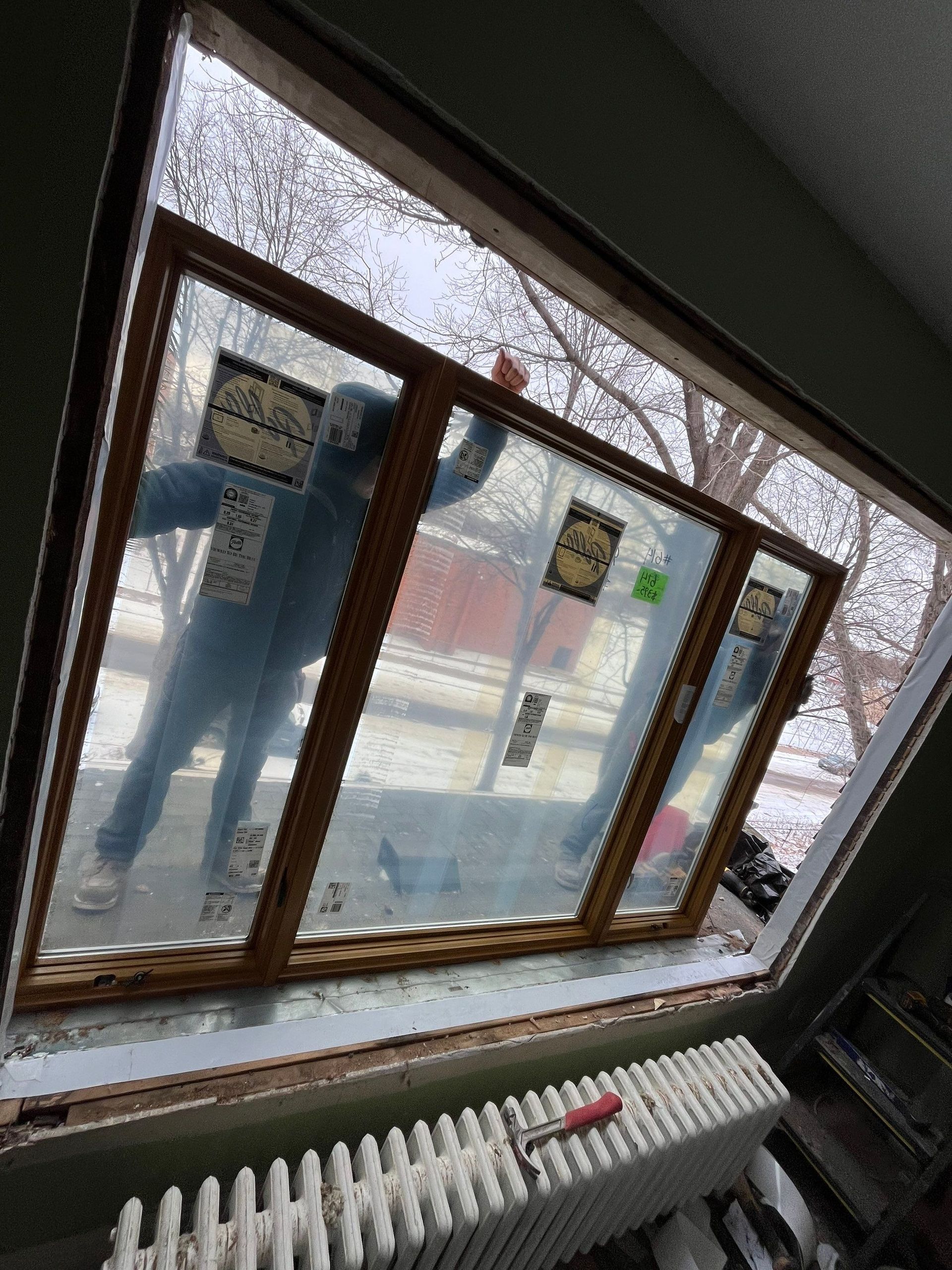 Person in blue holding new window inside a home, ready for installation; street view visible through glass.
