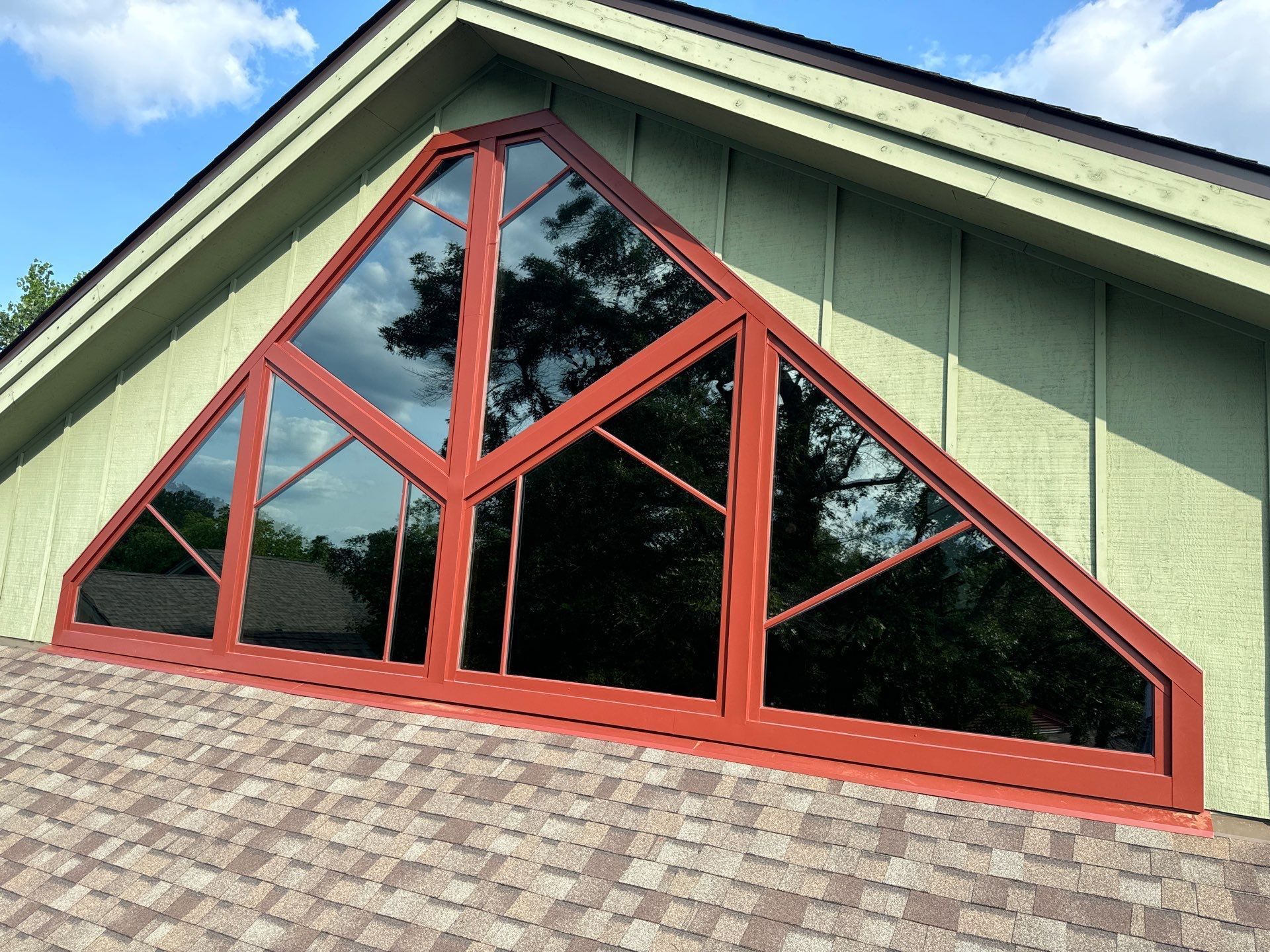 Red-framed triangular window on a green house with a brown roof, reflecting trees against a blue sky.