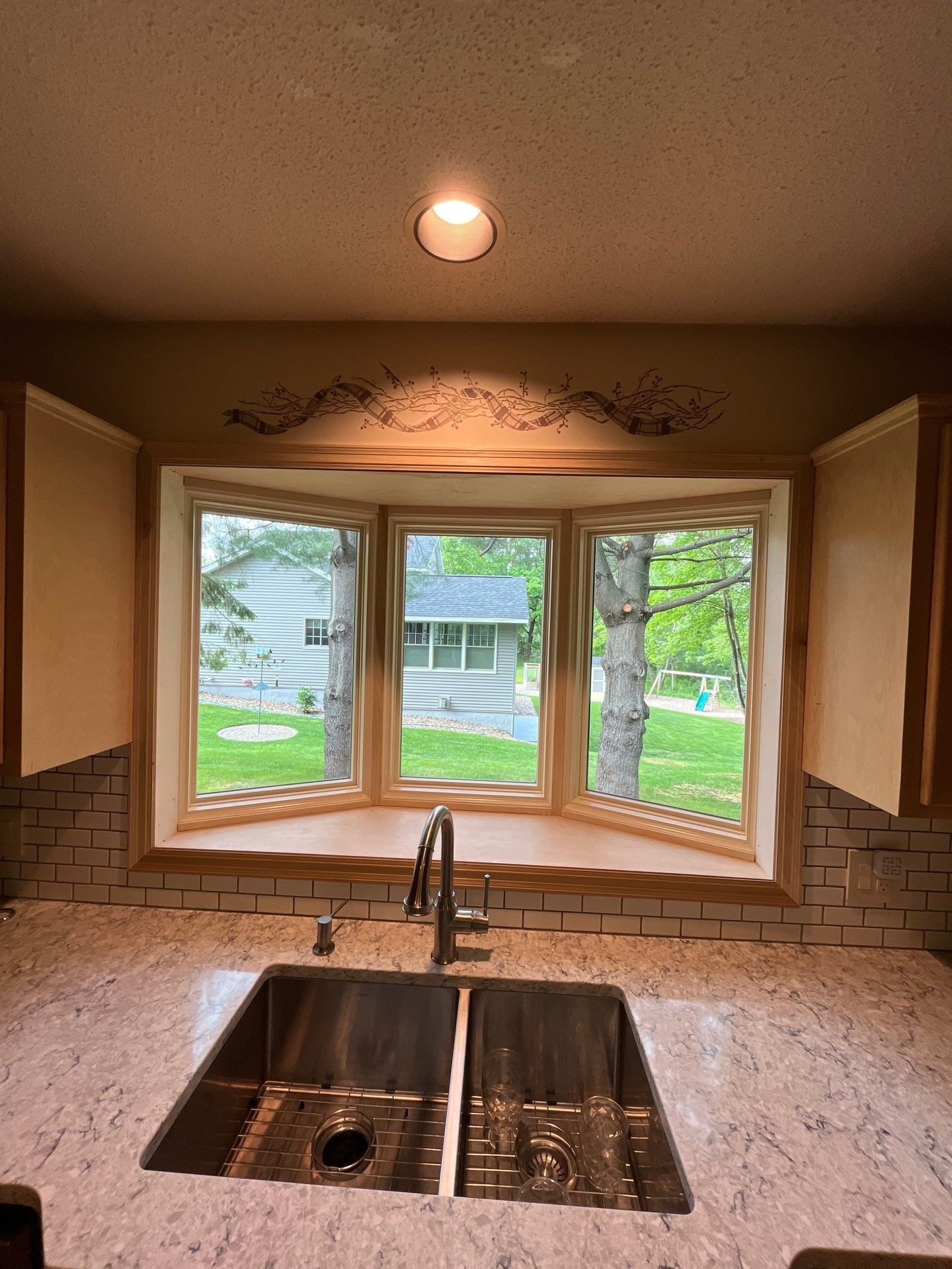 Kitchen with stainless steel sink under a bay window overlooking a green yard. Beige cabinets and white countertop.