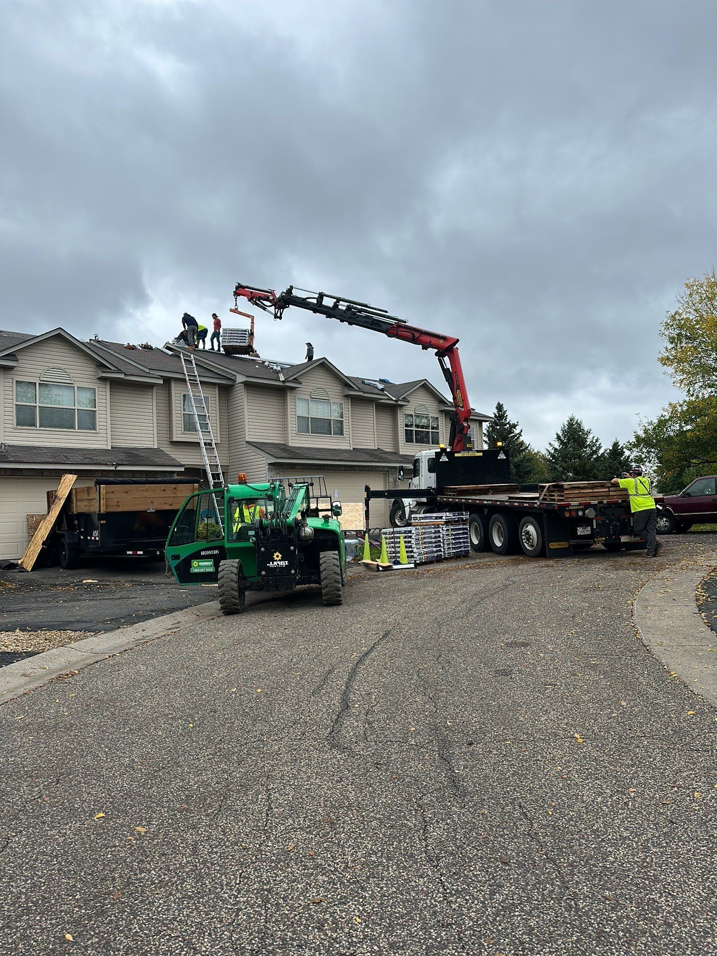 Roofers working on a house roof with a crane, truck, and tractor on a gravel driveway under an overcast sky.