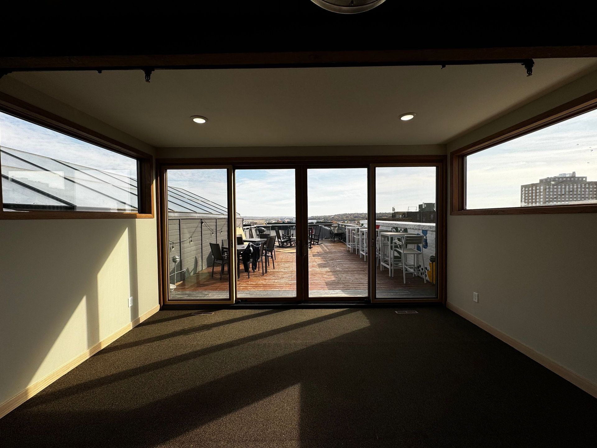 Room with large windows overlooking a rooftop deck with people and furniture; sunlight streams in.