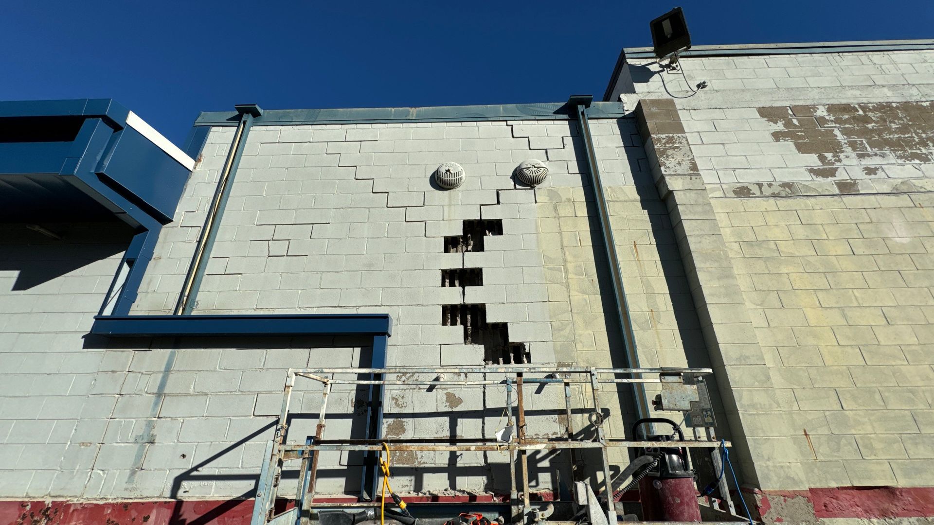 Damaged exterior brick wall with cracks, blue sky backdrop. A lift sits in front.