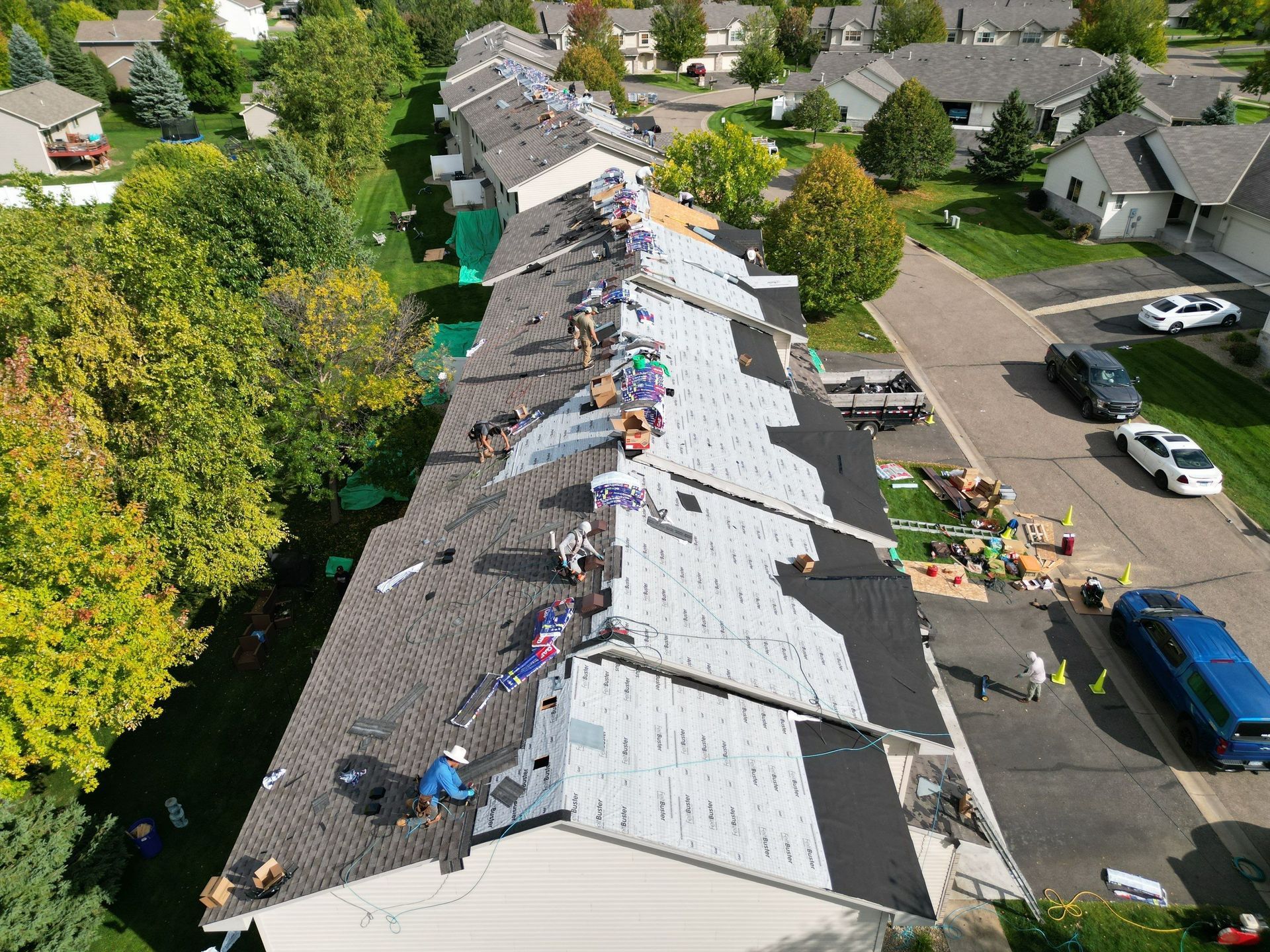 Roofers replacing shingles on a row of townhomes; various colors, equipment and cars visible.