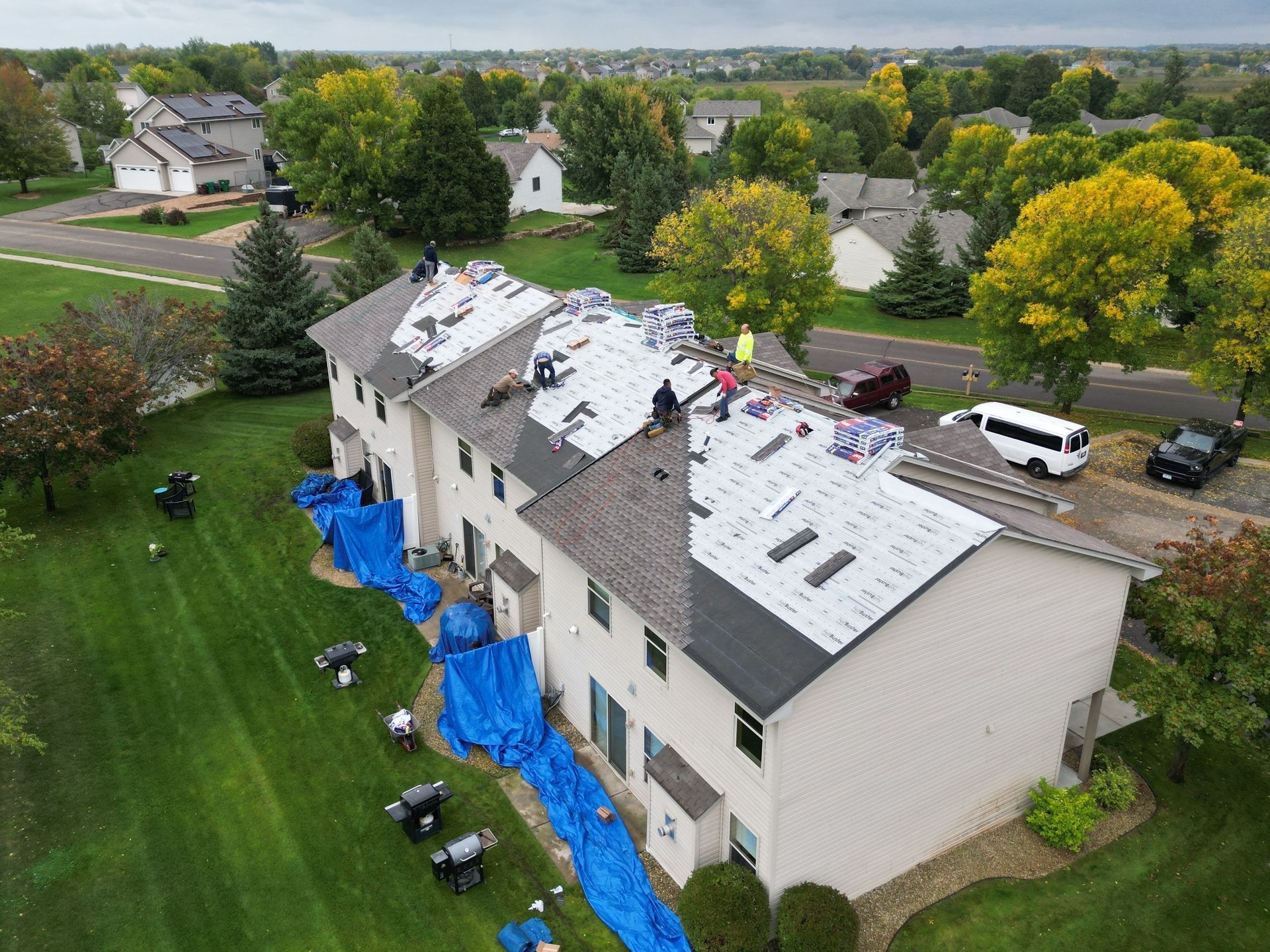 Roofers working on a multi-unit building, partially covered with blue tarps, surrounded by green grass and trees.