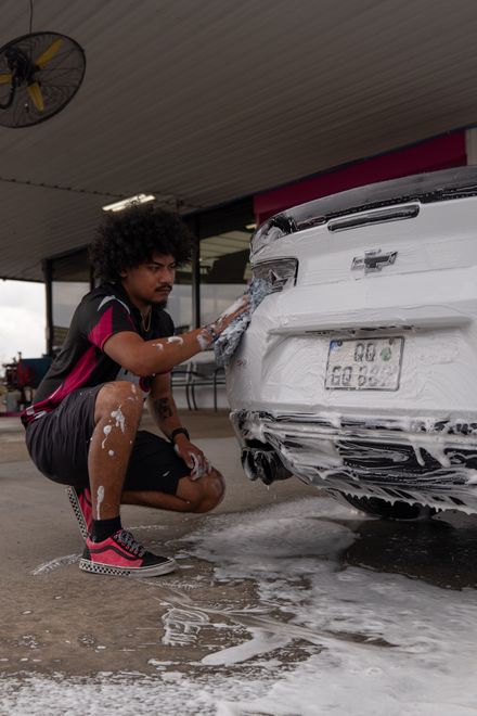 Man washing a white car covered in soap at an outdoor car wash, crouching and using a sponge.