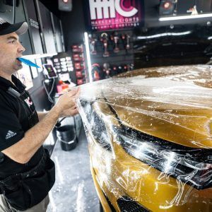 A man is covering a yellow car with plastic wrap.