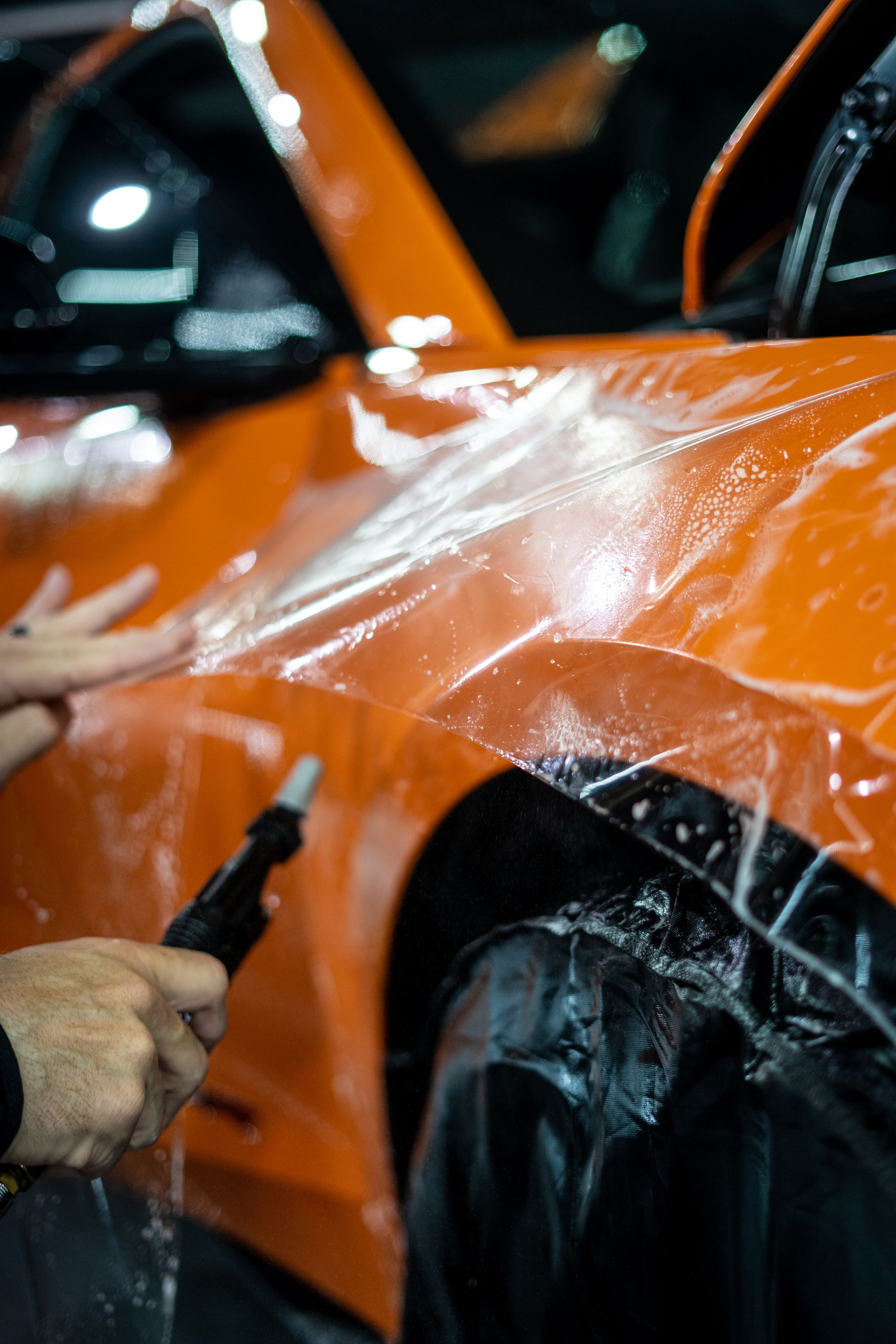 A person is applying a protective film to the side of an orange car.