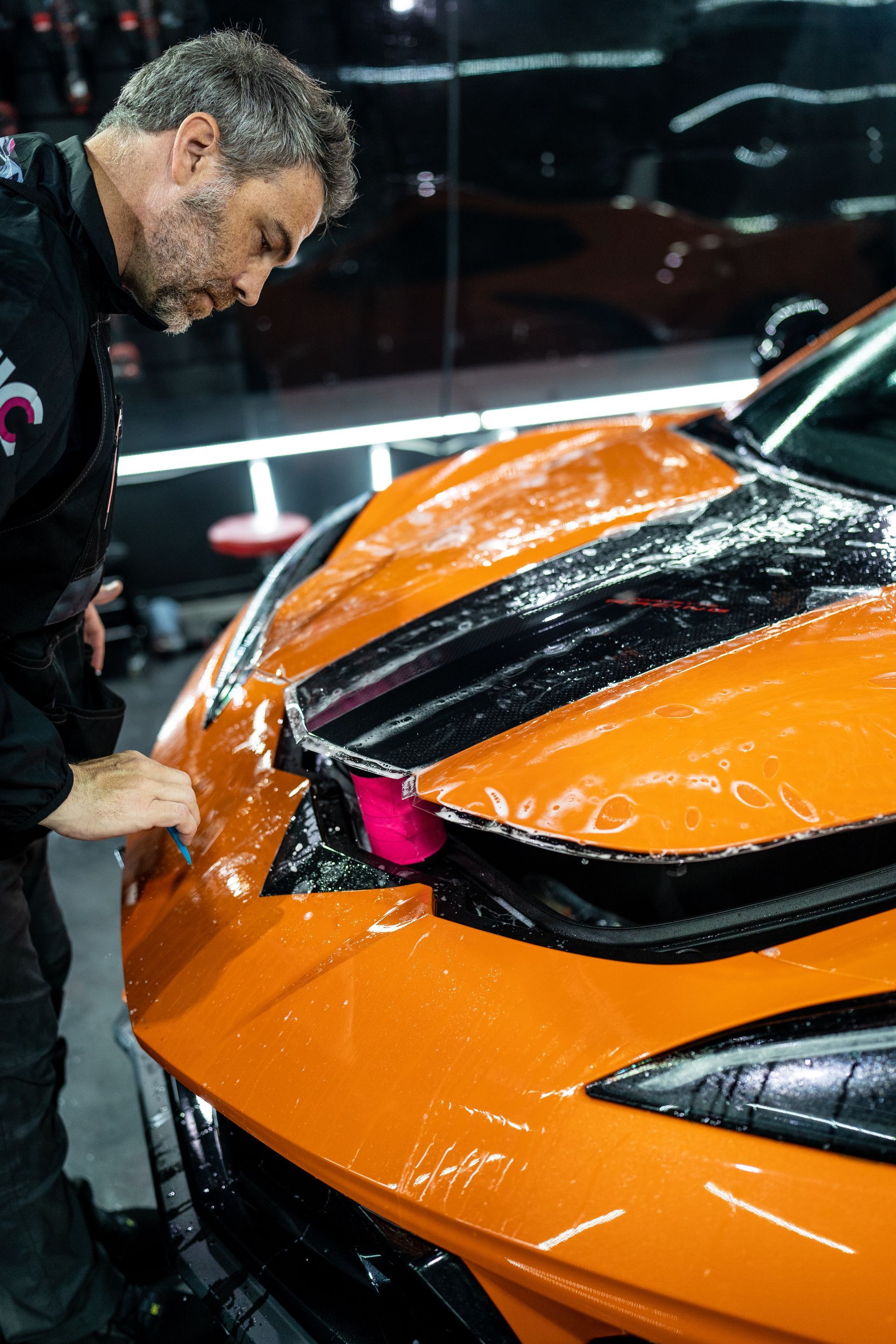 A man is applying a protective film to the front of an orange sports car.