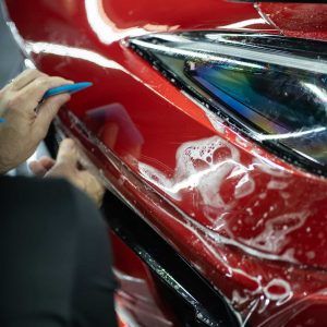 A person is applying a protective film to the front of a red car.