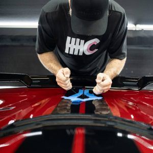 A man wearing a hhc shirt is polishing the hood of a red car.