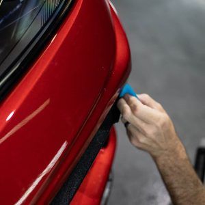 A person is polishing the front of a red car with a blue sponge.