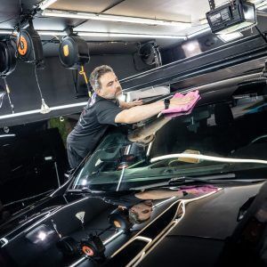 A man is cleaning the windshield of a black car in a garage.
