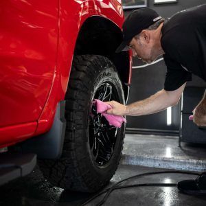 A man is cleaning the wheel of a red truck with a pink cloth.