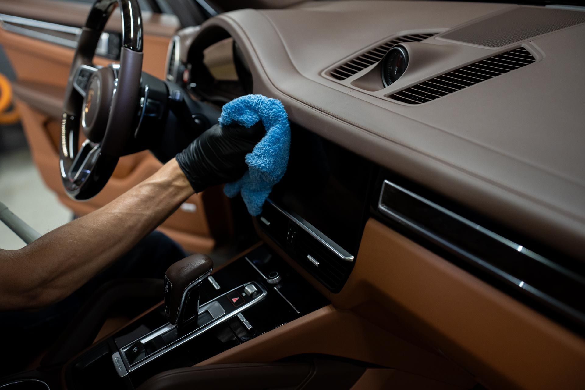 Person in black gloves cleaning a car dashboard with a blue cloth. The interior is tan leather.