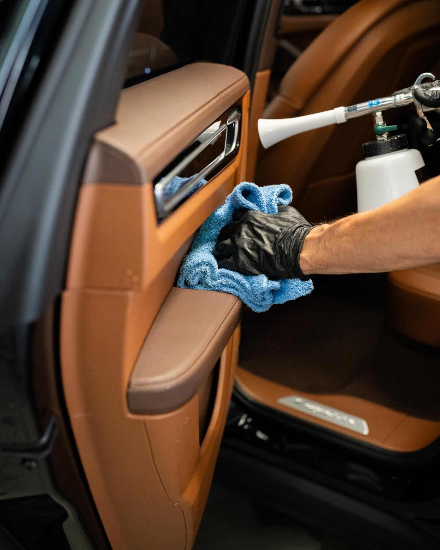 Person in black gloves cleaning car's tan door panel with a blue cloth and detailing gun.