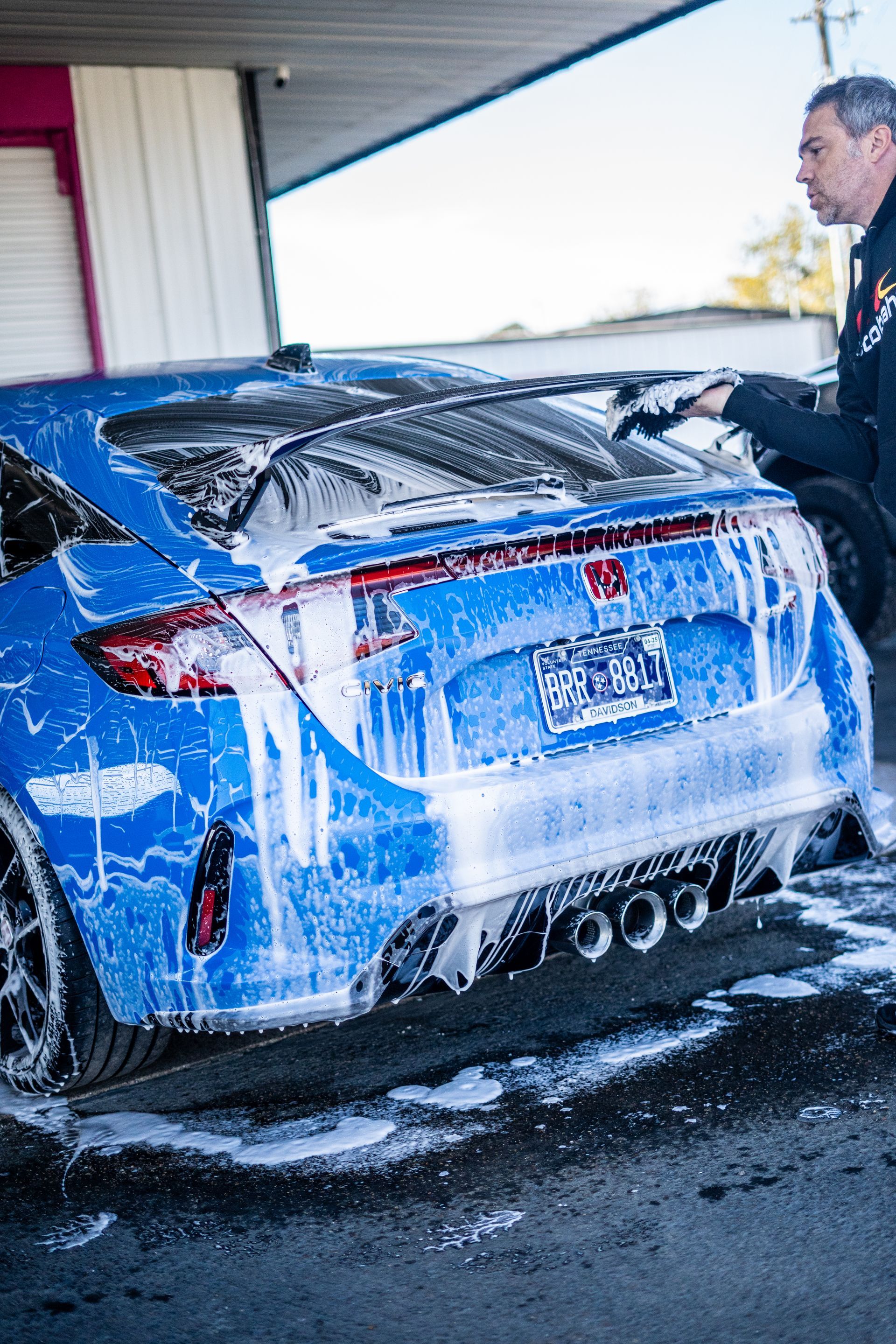 A blue Honda Civic being washed by a man at a car wash, covered in foam.